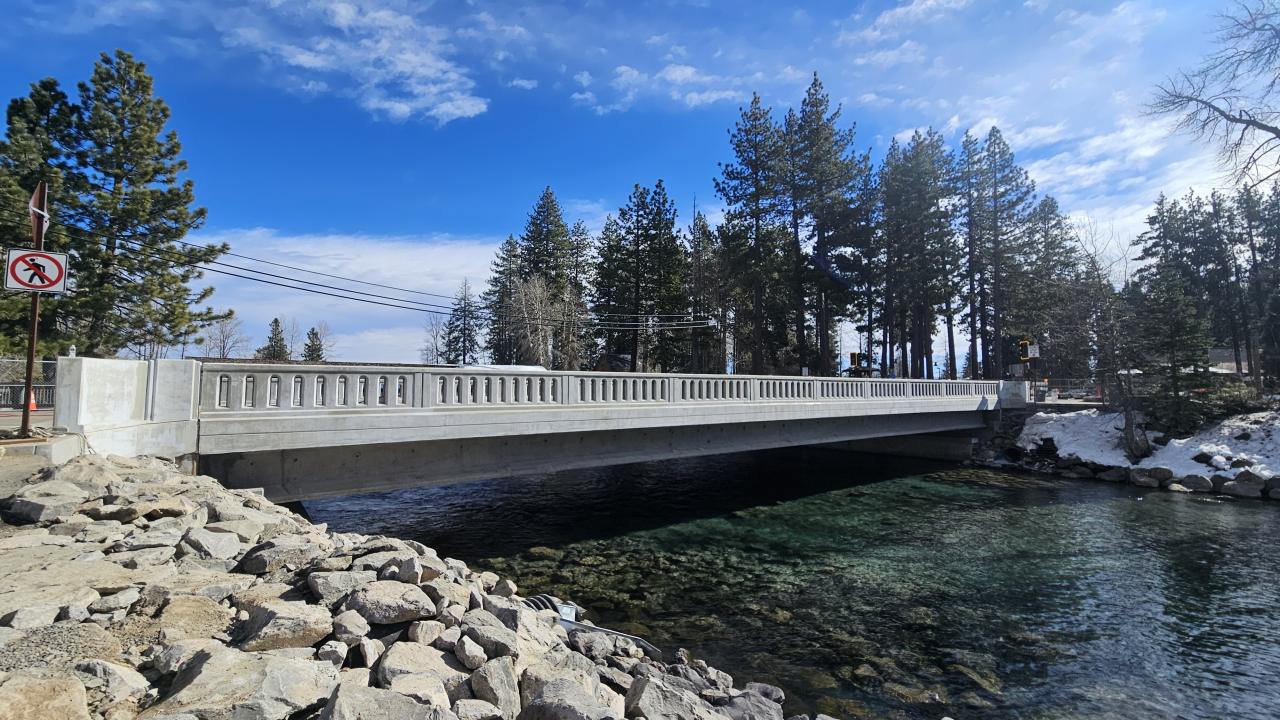 Concrete bridge over a clear stream, surrounded by pine trees and a blue sky.
