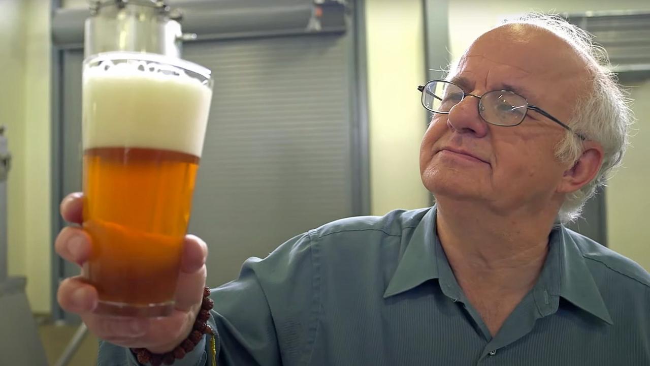 An older man wearing glasses examines a glass of beer in a brewery setting.