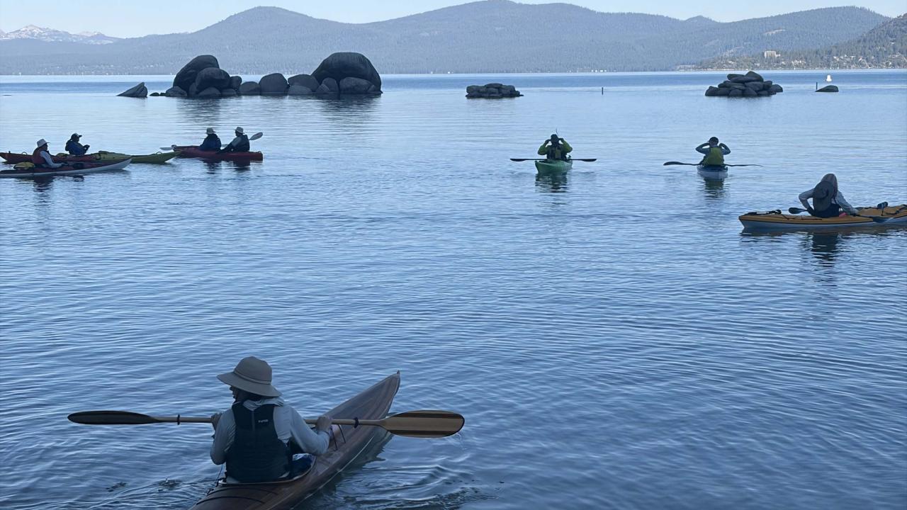 Kayakers paddling on a calm blue lake; orange inflatable tube on sandy shore, mountains beyond.
