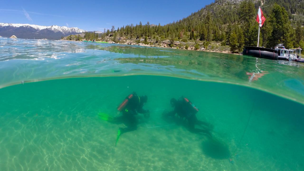 Split-view lake: two scuba divers in clear turquoise water; forested mountains and dock above.