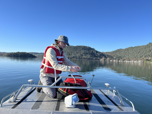 Person in life jacket and bucket hat kneeling on small boat adjusting a red cooler on calm lake