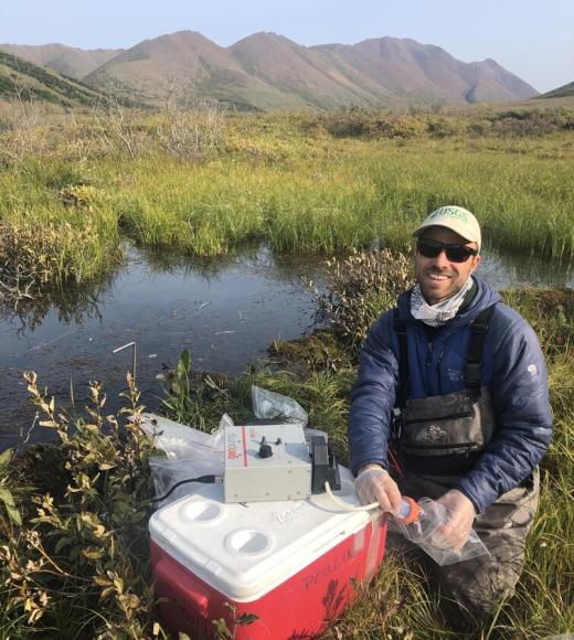 Man kneeling by red cooler at grassy riverside with mountains in background
