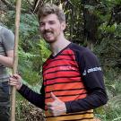 Smiling man giving thumbs up holding a stick in forest, wearing red-striped jersey