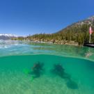 Split-view lake: two scuba divers in clear turquoise water; forested mountains and dock above.