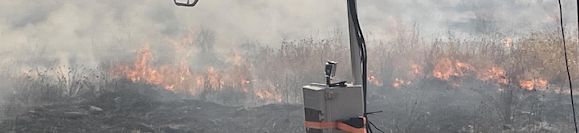 Sensor mast with camera in foreground, grass wildfire and smoke burning behind